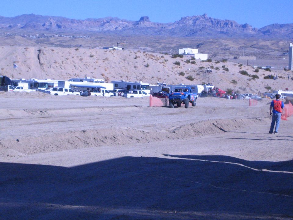 Blue off-road race truck on a desert dirt course past parked RVs and trucks