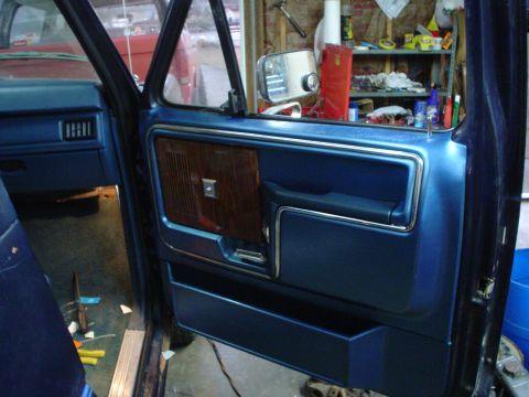 1984 Ford Bronco - Interior of an open blue pickup door showing the door panel with woodgrain insert and chrome trim inside a cluttered garage.