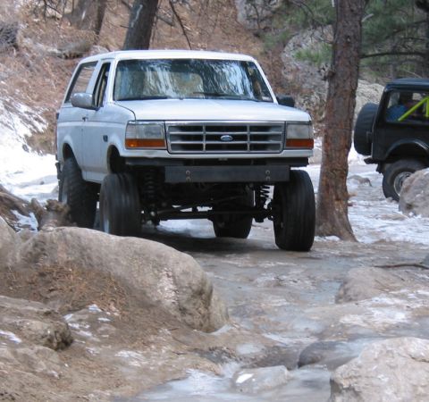 1981 Ford Bronco - A lifted white Ford Bronco navigates a challenging off-road course with icy patches and large rocks.
