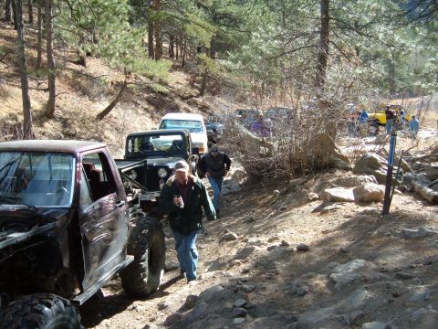 1993 Ford Bronco - A convoy of off-road vehicles, including a 1993 Ford Bronco, navigates a challenging rocky trail through a wooded area.