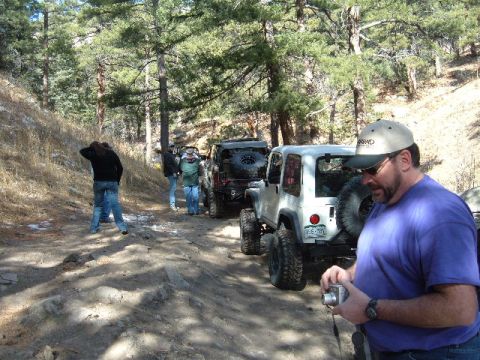1993 Ford Bronco - A group of Jeeps are lined up on a dirt path surrounded by trees, with a man holding a camera in the foreground.