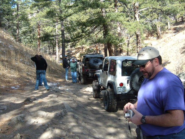 Several Jeeps parked on a dirt road in a wooded area. A man in a purple shirt holds a camera