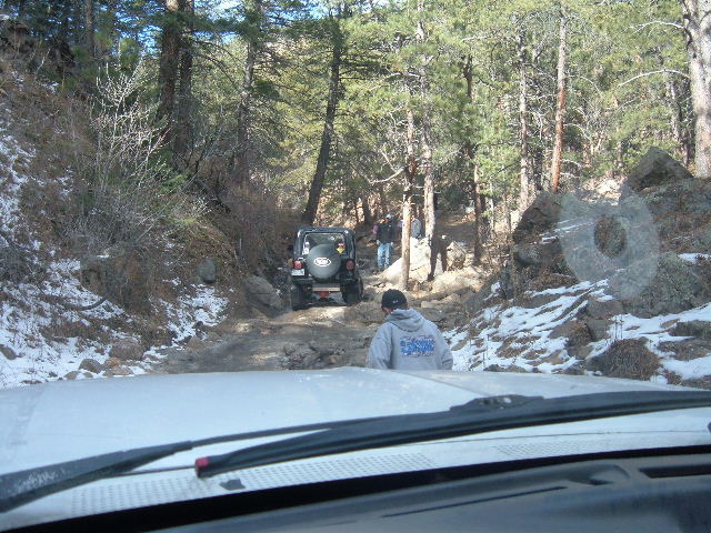 Black Jeep Wrangler on a rocky, snow-dusted trail with trees and people