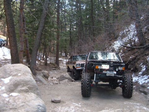 1993 Ford Bronco - Two modified off-road vehicles navigate a wooded trail with snow patches on the ground.