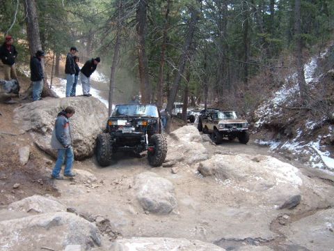 1993 Ford Bronco - Two off-road vehicles navigate a rocky, wooded trail with snow patches visible.