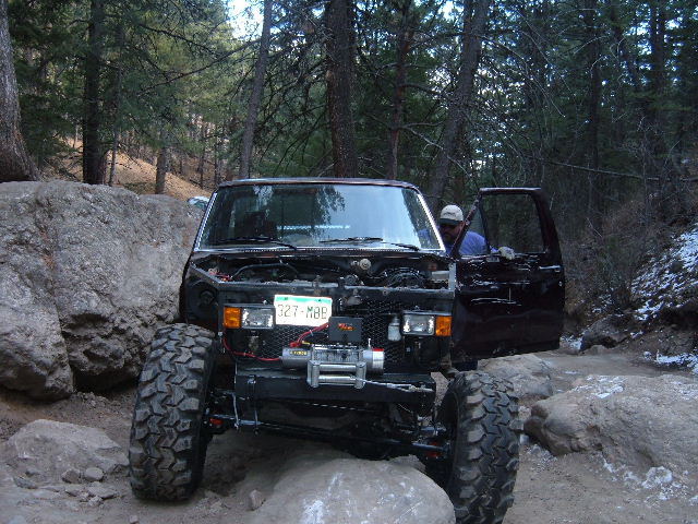 Black 1993 Ford Bronco off-roading, hood open, winch mounted, with large tires navigating rocks