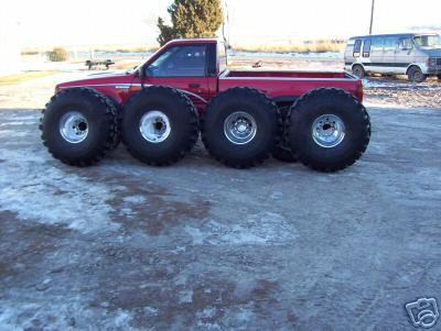 1979 Ford Bronco - A red pickup truck is modified with six enormous tires, parked next to a white van.
