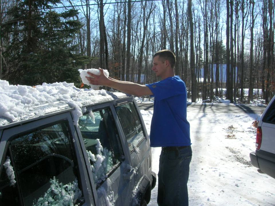 Man in blue shirt removing snow from the roof of a dark colored SUV. Another white SUV is parked to the right