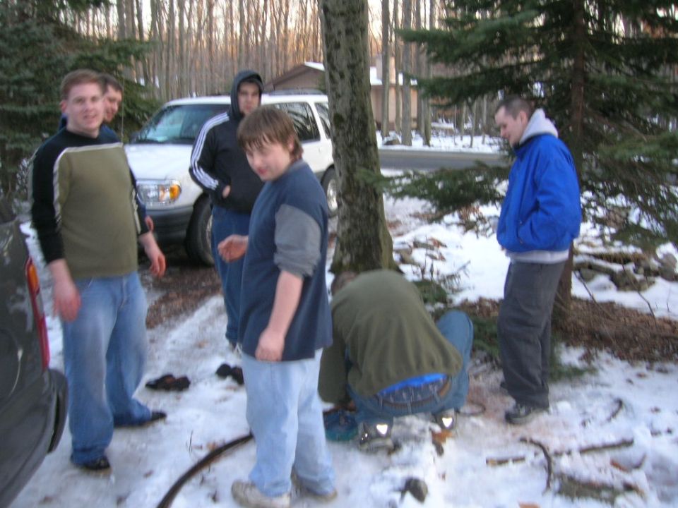 Group of guys standing around a white SUV in a snowy wooded area. One person is working on something on the ground