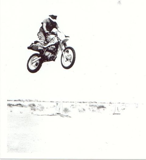 1900 Honda Big Red - A dirt bike rider soars through the air, performing a jump over a barren landscape.