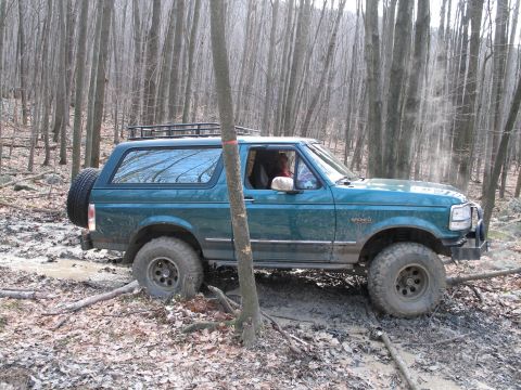 1995 Ford Bronco - A teal 1995 Ford Bronco navigates a muddy path in a wooded area, kicking up dirt.