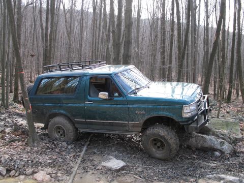 1995 Ford Bronco - A green Ford Bronco navigates a muddy, wooded trail with large tires and a roof rack.