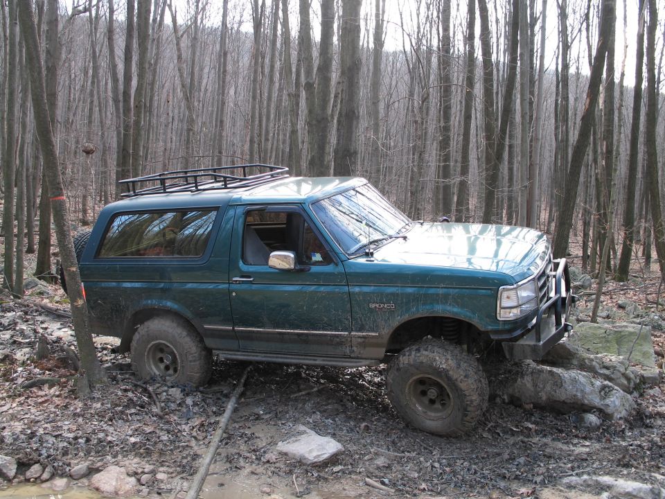 Green Ford Bronco with a roof rack and bull bar, driving off-road in a wooded area