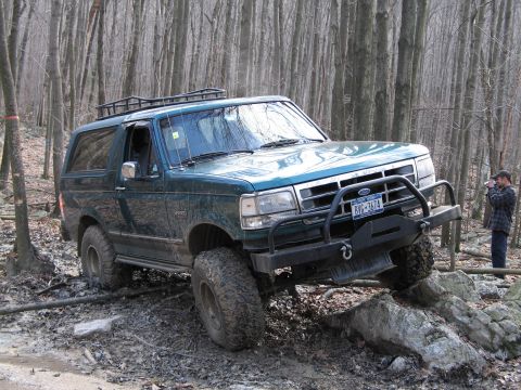 1995 Ford Bronco - A green 1995 Ford Bronco tackles a challenging off-road course in a wooded area.