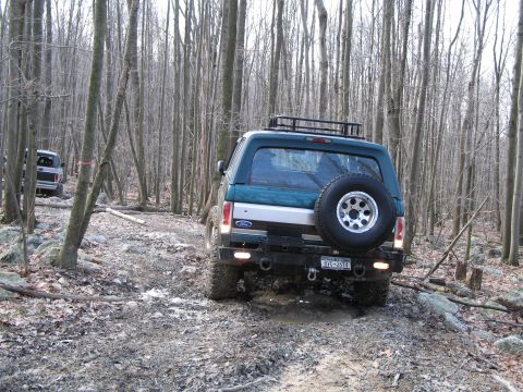 1995 Ford Bronco - Two SUVs navigate a rocky, wooded path. The lead vehicle is a 1995 Ford Bronco.