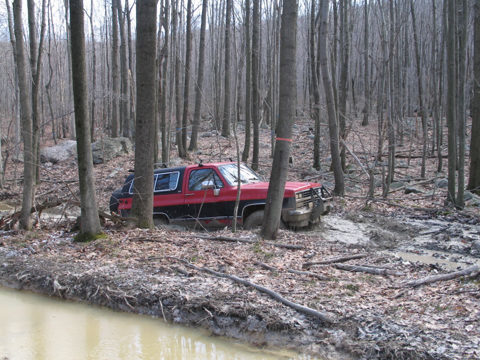 Red and black SUV with off-road bumper stuck in mud, surrounded by trees