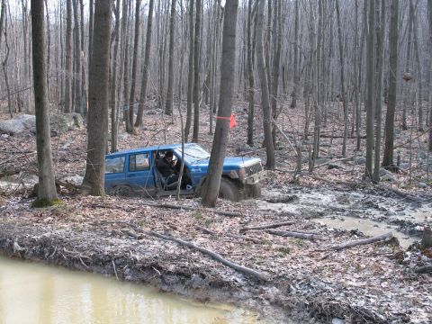 1995 Ford Bronco - A blue Jeep Cherokee navigates a muddy trail through a dense forest.