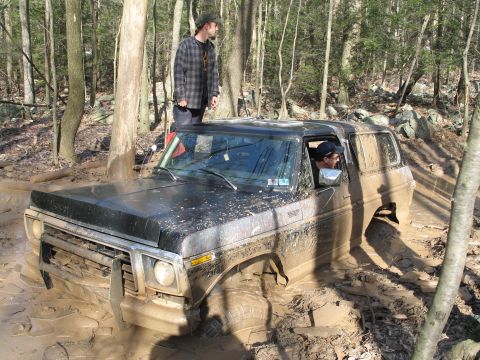 1995 Ford Bronco - A black SUV is stuck deep in mud on a wooded trail, with two people visible.