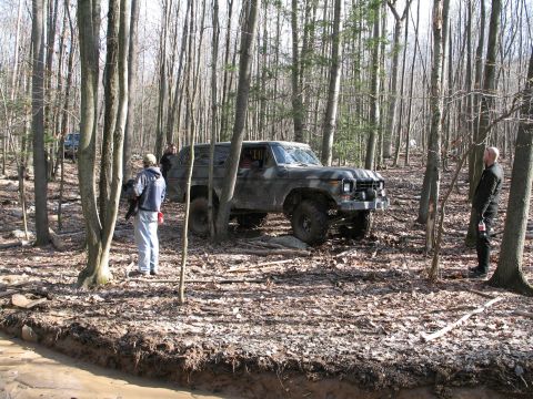 1995 Ford Bronco - A group of people are gathered around an off-road vehicle in a wooded area with muddy terrain.