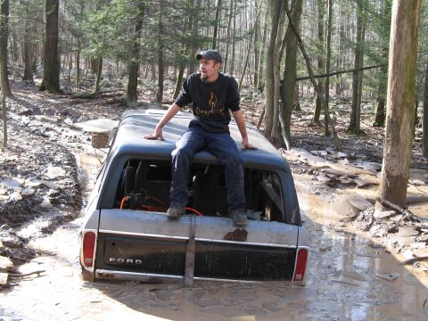 1995 Ford Bronco - A man sits atop a mud-covered Ford Bronco, which is partially submerged in a wooded area.