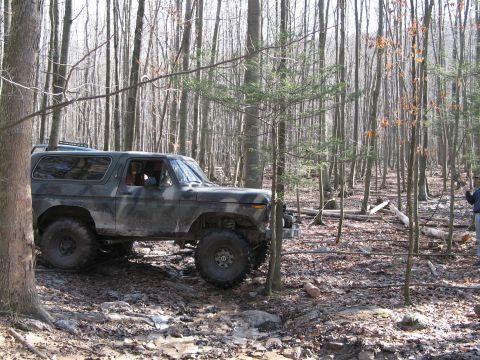 1995 Ford Bronco - A camouflaged Ford Bronco tackles a challenging muddy path through a dense forest.
