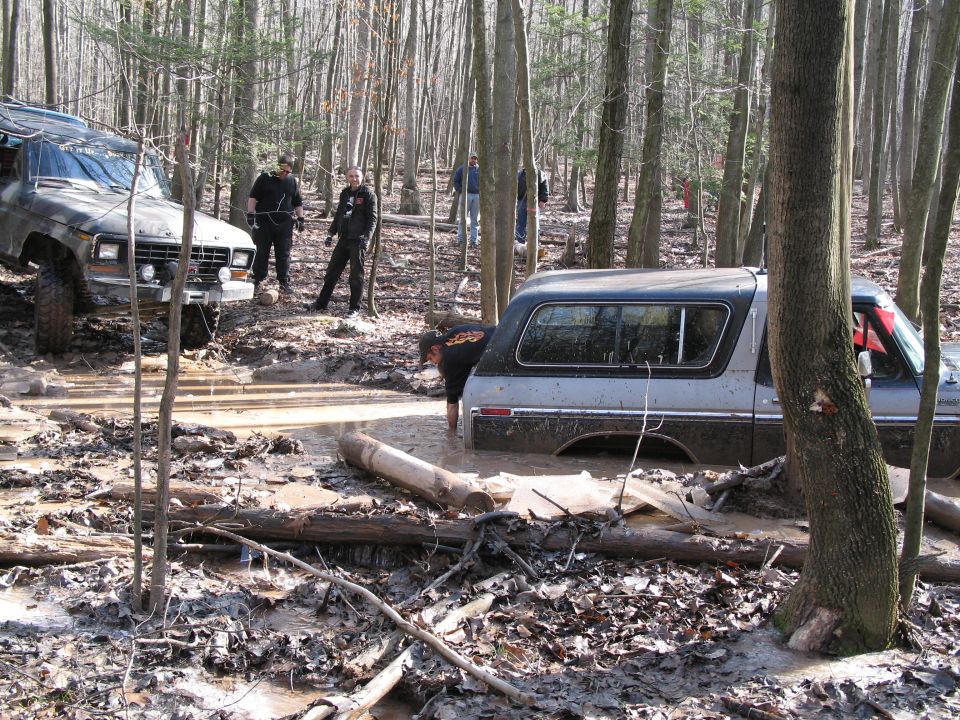 two off-road vehicles stuck in mud in a wooded area, one is a 1978 Ford Bronco