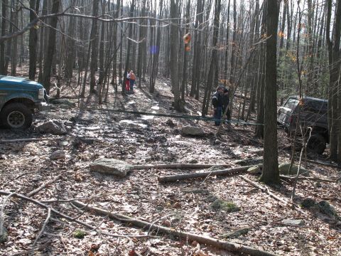 1995 Ford Bronco - Two off-road vehicles navigate a muddy, wooded trail with fallen branches and rocks.