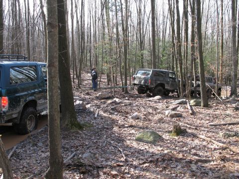 1995 Ford Bronco - Two SUVs navigate a wooded area with muddy terrain and scattered rocks.