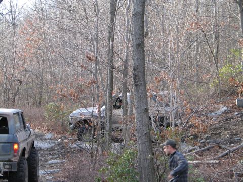 1995 Ford Bronco - Two modified SUVs navigate a wooded trail with fallen leaves on the ground.