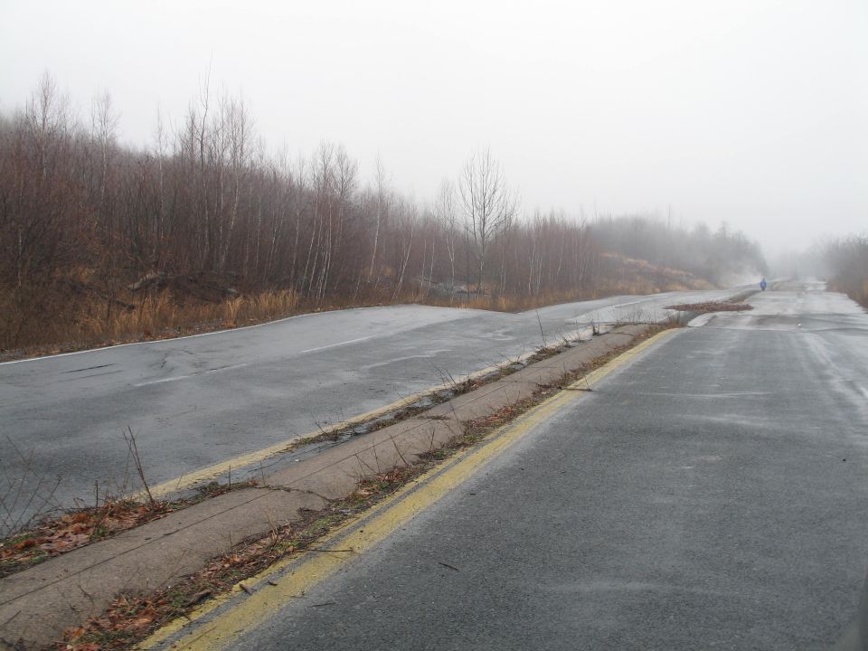 Damaged asphalt road with yellow lines, overgrown with weeds, leading into a foggy landscape with bare trees
