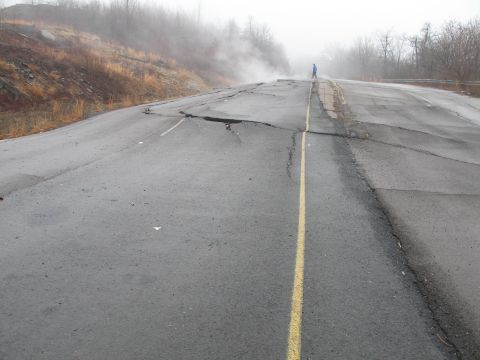 1995 Ford Bronco - A person stands on a severely damaged road with large cracks and buckling asphalt, indicating a significant structural failure.