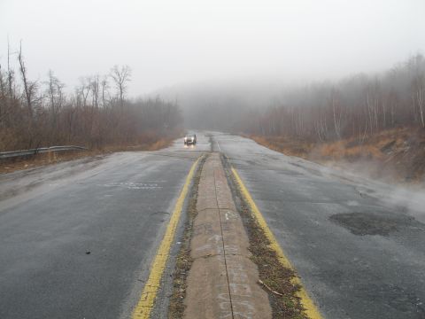 1995 Ford Bronco - A single car drives down a wet, fog-shrouded road surrounded by bare trees.