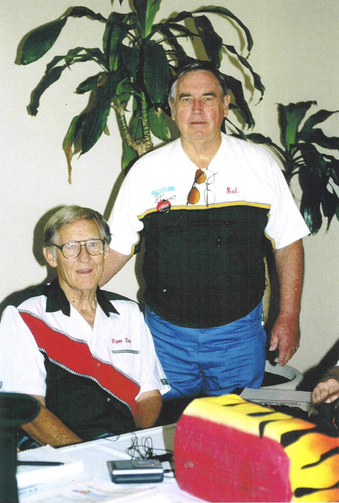 Two men indoors, one seated wearing a black, white, and red shirt, the other standing behind him wearing a black and white shirt with sunglasses on the collar