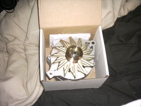 1991 Ford LTD Crown Victoria - A new alternator sitting fan-up in an open white cardboard box, shown from above with its gold-tinted cooling fan blades, pulley nut and ca…