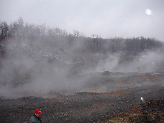 Person wearing a red baseball cap stands in a misty, rocky landscape with bare trees in the background