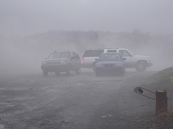 Tan Jeep Grand Cherokee, white SUV and blue sedan parked in dense fog on gravel