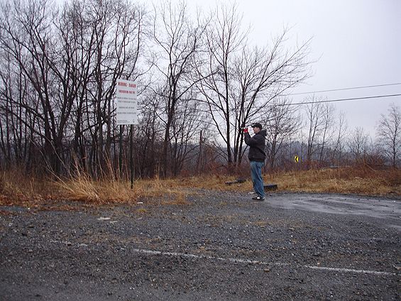 Person photographing from a gravel lot beside a warning sign and bare winter trees