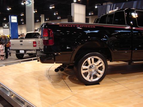 2000 Ford Crown Victoria - A black pickup truck with Harley-Davidson branding is displayed at an auto show alongside another truck.