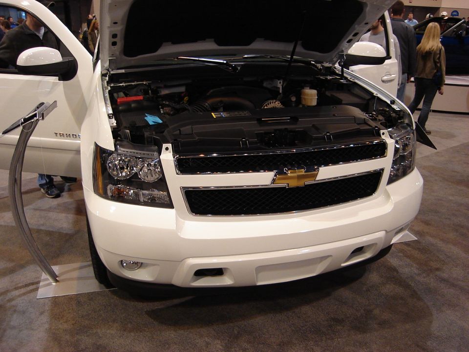 Front view of a white Chevrolet SUV, hood open, showing the engine bay. The Chevrolet logo is prominent on the grille
