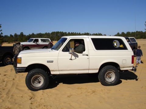 1989 Ford Bronco - A white 1989 Ford Bronco is parked on sand with other vehicles in the background.