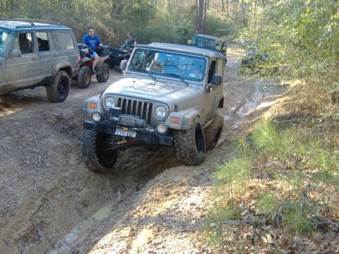 1999 Jeep Wrangler - Multiple off-road vehicles navigate a muddy trail through a wooded area.