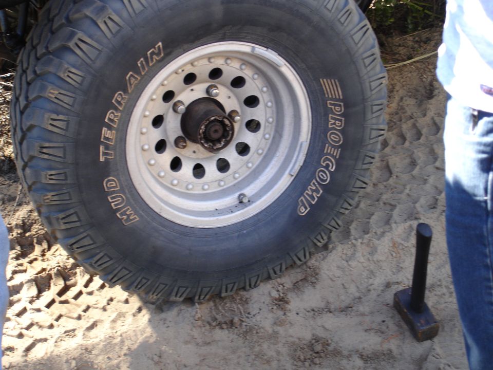 Close-up of a large off-road tire with 'MUD TERRAIN' and 'PROCOMP' text, mounted on a silver wheel. Tire tracks visible in the sand