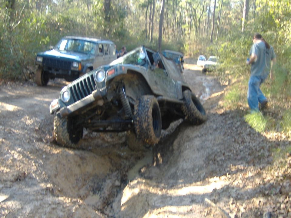 Jeep Wrangler going up a muddy hill with another Jeep Cherokee behind it. Person in blue jeans and t-shirt watches