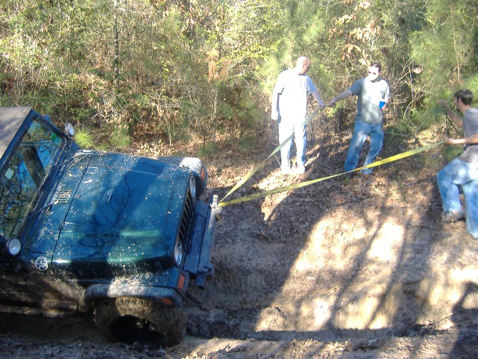 Dark green Jeep Wrangler stuck in mud, being pulled by a yellow strap by three people in a wooded area