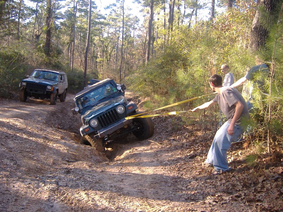 1999 Jeep Wrangler stuck in a muddy ditch, being pulled by a yellow tow strap attached to another Jeep off-road