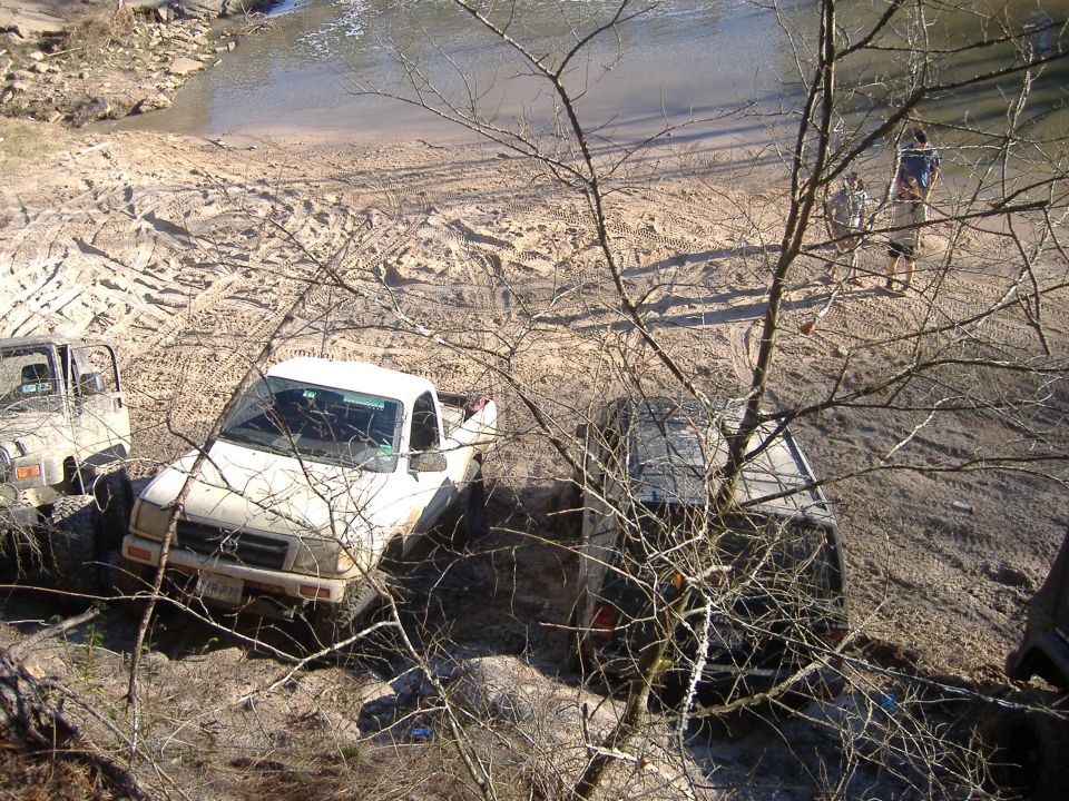 Muddy white pickup truck and Jeep Wrangler stuck in dirt near water, with two people standing in background