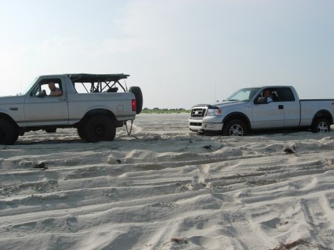 1995 Ford Bronco - Two trucks, a Ford Bronco and a Ford F-150, are stuck in the sand on a beach.