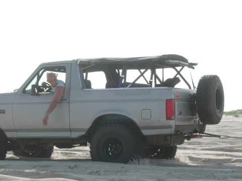 1995 Ford Bronco - Man driving a 1995 Ford Bronco on a sandy beach, spare tire visible on the back.