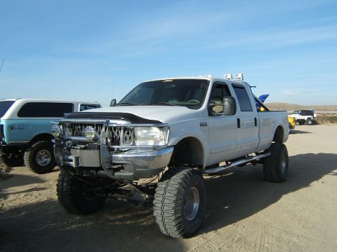 1988 Ford Bronco - A lifted white pickup truck is parked on a dirt lot under a clear blue sky, with other vehicles visible in the distance.