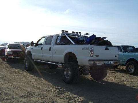 1988 Ford Bronco - A white pickup truck with off-road tires and a roll bar is parked on sandy terrain next to other vehicles.